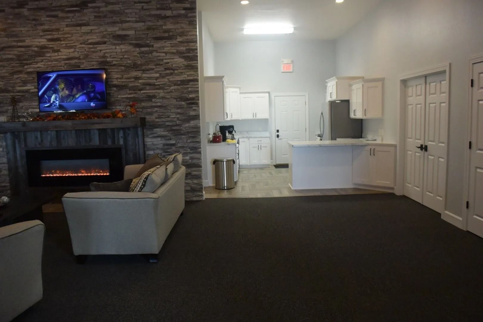 Spacious lounge with sofa, fireplace, wall-mounted TV, and kitchenette area in the back.