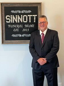 Man in dark suit standing beside a sign reading “Sinnott Funeral Home Est 2015”