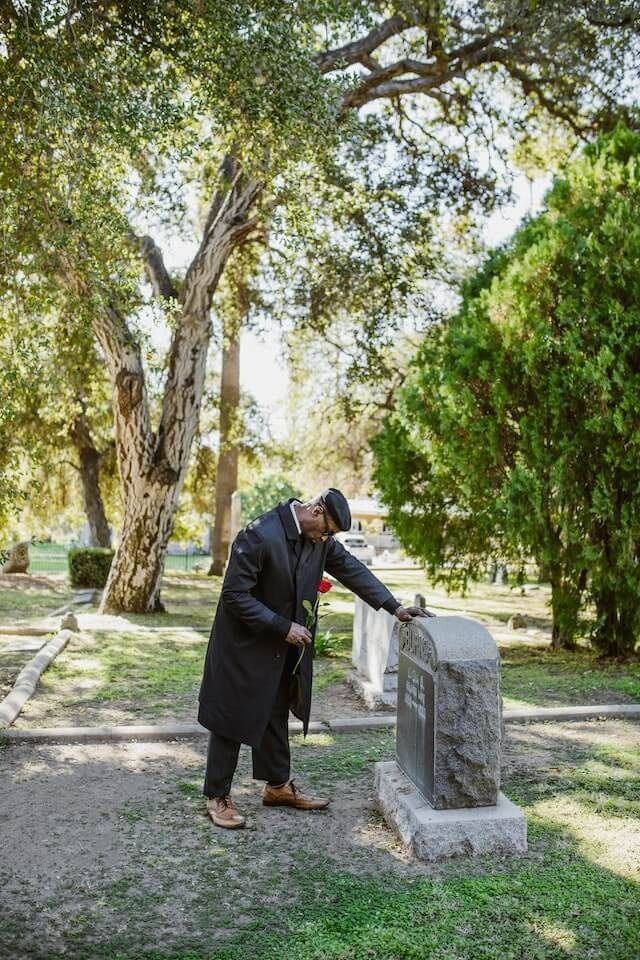 Person in a black coat leans over a gravestone in a tree-lined cemetery or park.
