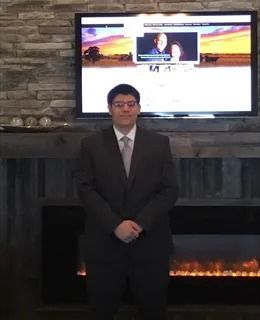 Man in a dark suit standing indoors in front of a fireplace and wall-mounted TV.
