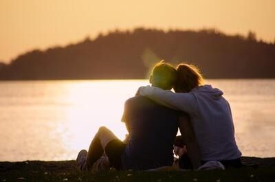 Two people sitting by a lake at sunset, embracing with a glowing sky behind them.