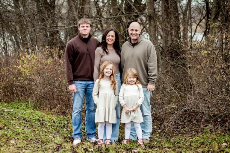 Family of four standing outdoors in a wooded area, smiling at the camera.