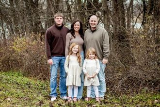 Family of four standing outdoors in a wooded area, smiling at the camera.
