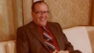 Smiling man in a brown suit and striped tie seated on a couch