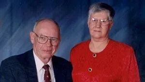 Two adults posed for a studio portrait against a blue backdrop, one in a dark suit and one in a red blazer.