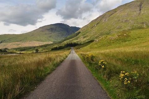 Narrow paved road through green hills and grassy fields under a cloudy sky.