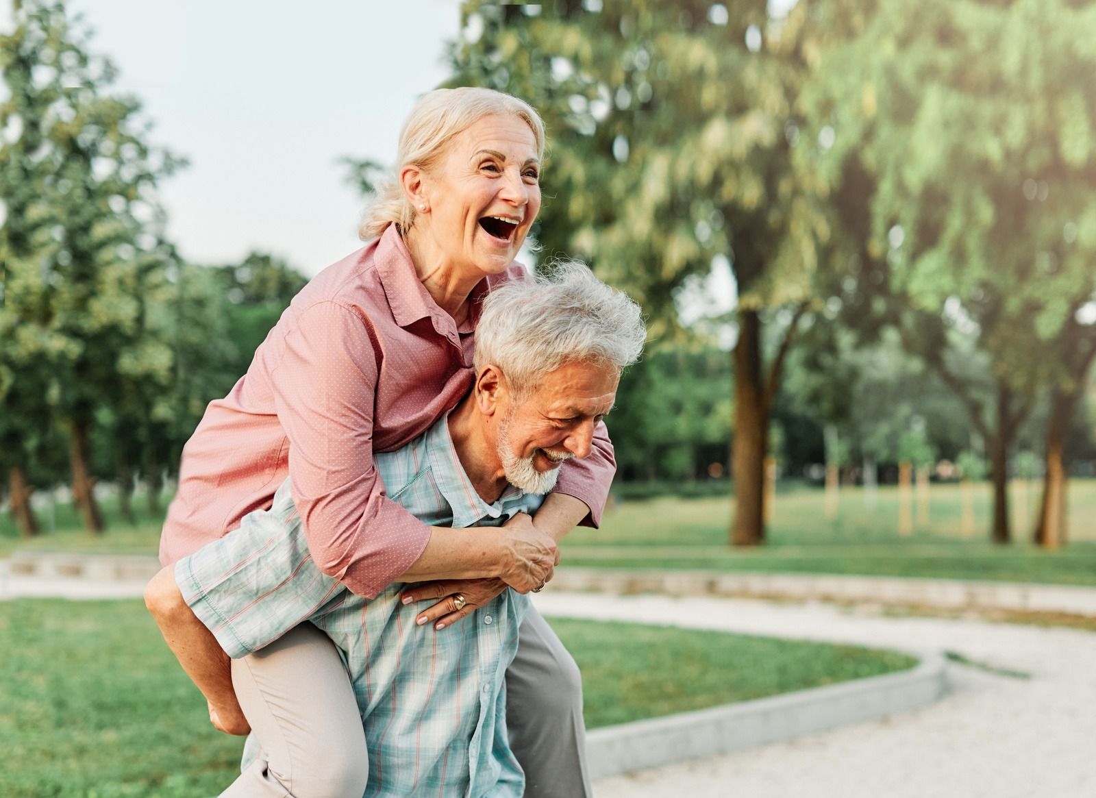 Smiling older woman piggybacks a smiling older man in a park with trees and a winding path.