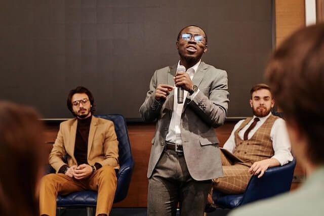 Man speaking into a microphone at a panel discussion, with two seated audience members behind him.