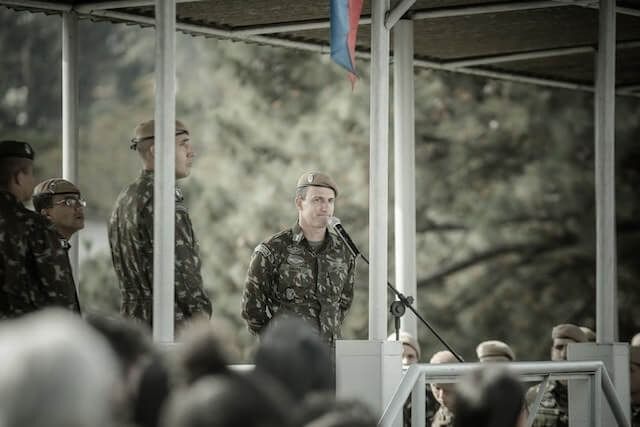 Soldiers in camouflage stand at attention on a covered platform during a public event