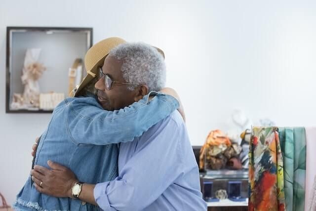 Two people embrace warmly in a bright room with artwork and clothing in the background.