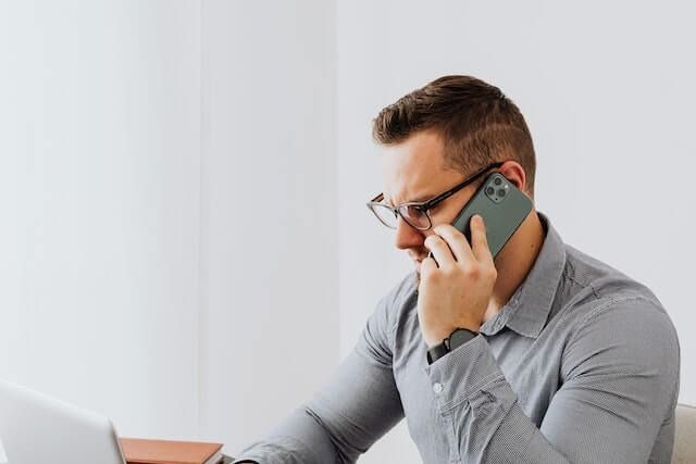 Man in glasses talking on a smartphone while working at a laptop in a bright office