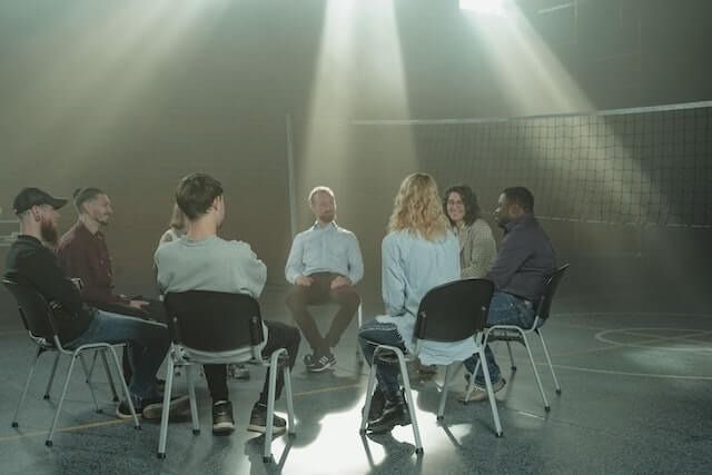 Seven people sit in a circle in a dim room with bright light streaming down from above.
