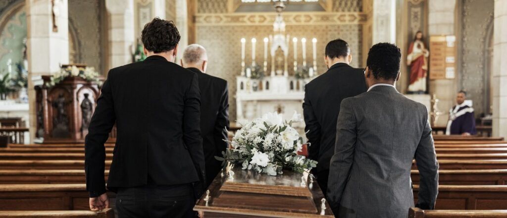 Mourners in dark suits stand in a church aisle facing a flower-covered altar and casket