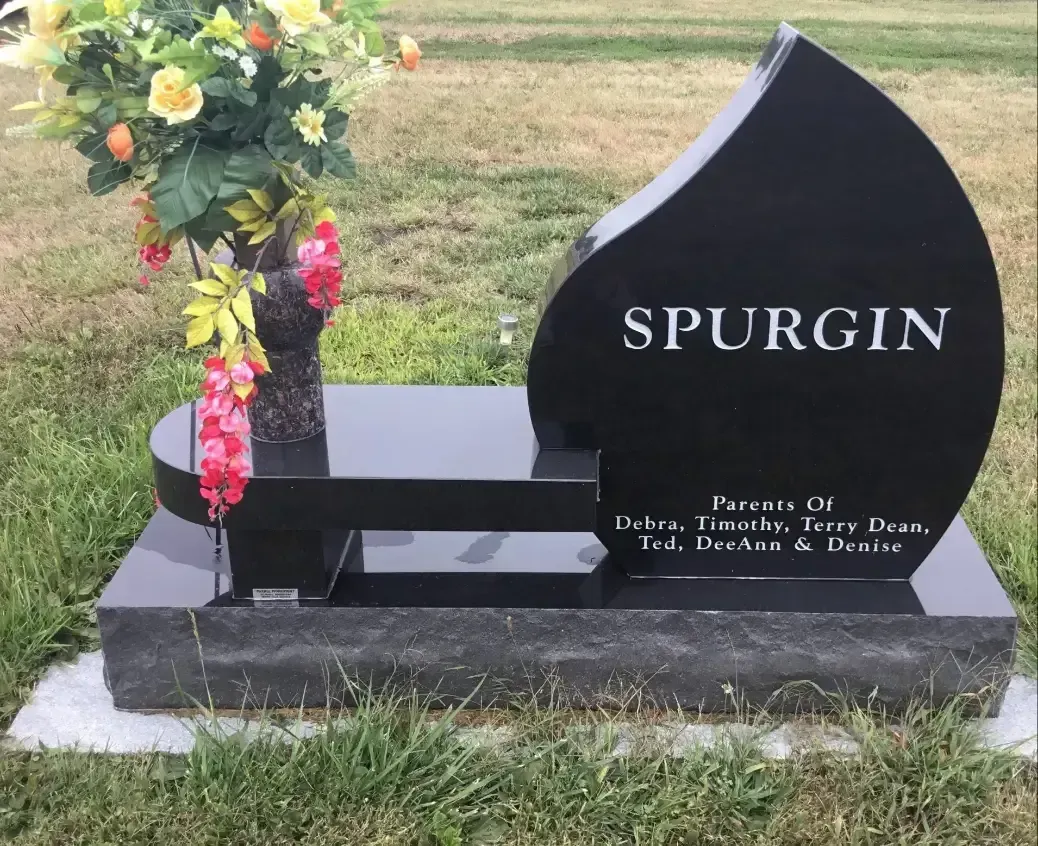 Black memorial headstone with “SPURGIN” and flower arrangement on a grassy grave site.