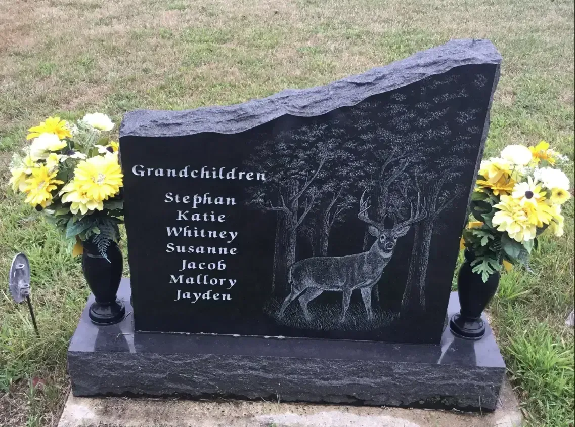 Granite memorial with engraved names and deer, flanked by yellow flowers on grass