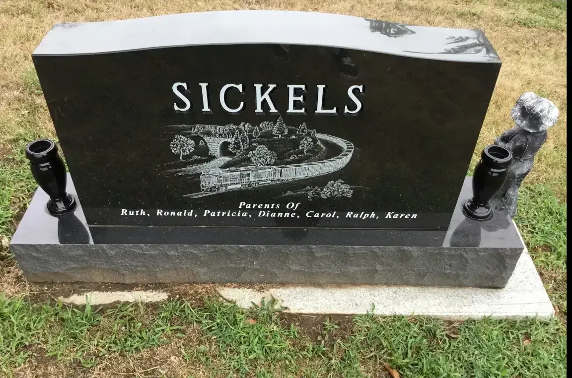 Black gravestone with “SICKELS” engraved, featuring decorative designs and small statues on a grassy plot.