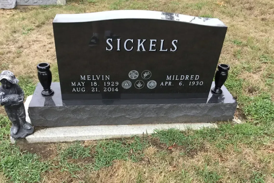 Black cemetery headstone with “Sickels” engraved, beside small memorial figurines on a grassy plot.