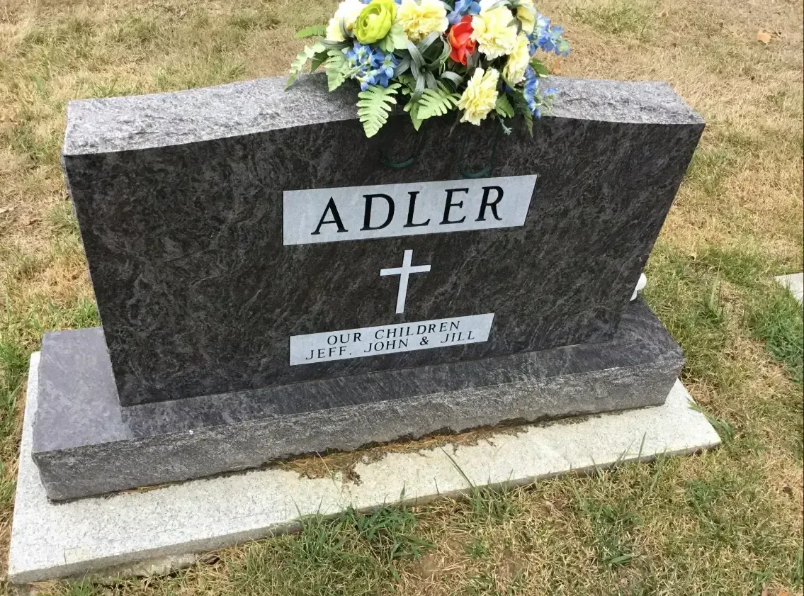 Gray granite gravestone with “ADLER,” a cross, and flowers on a grassy plot.