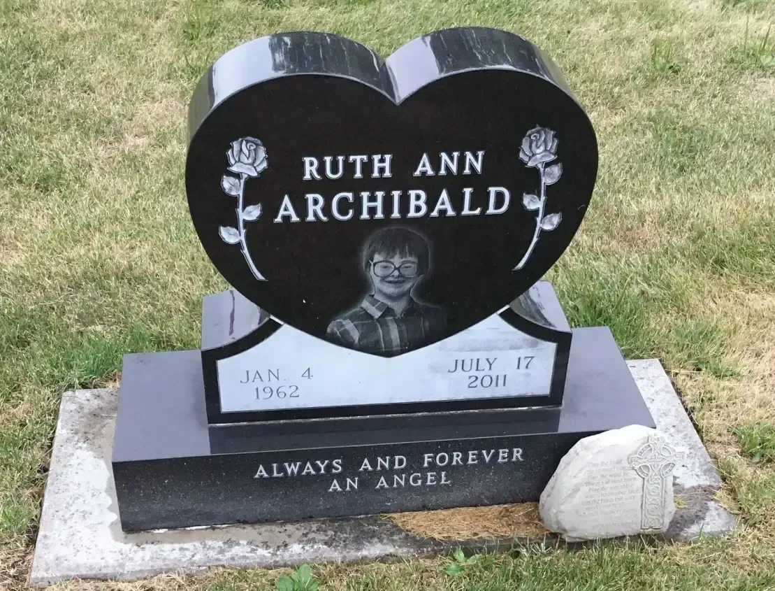 Black heart-shaped gravestone on grass with flowers, inscribed 
