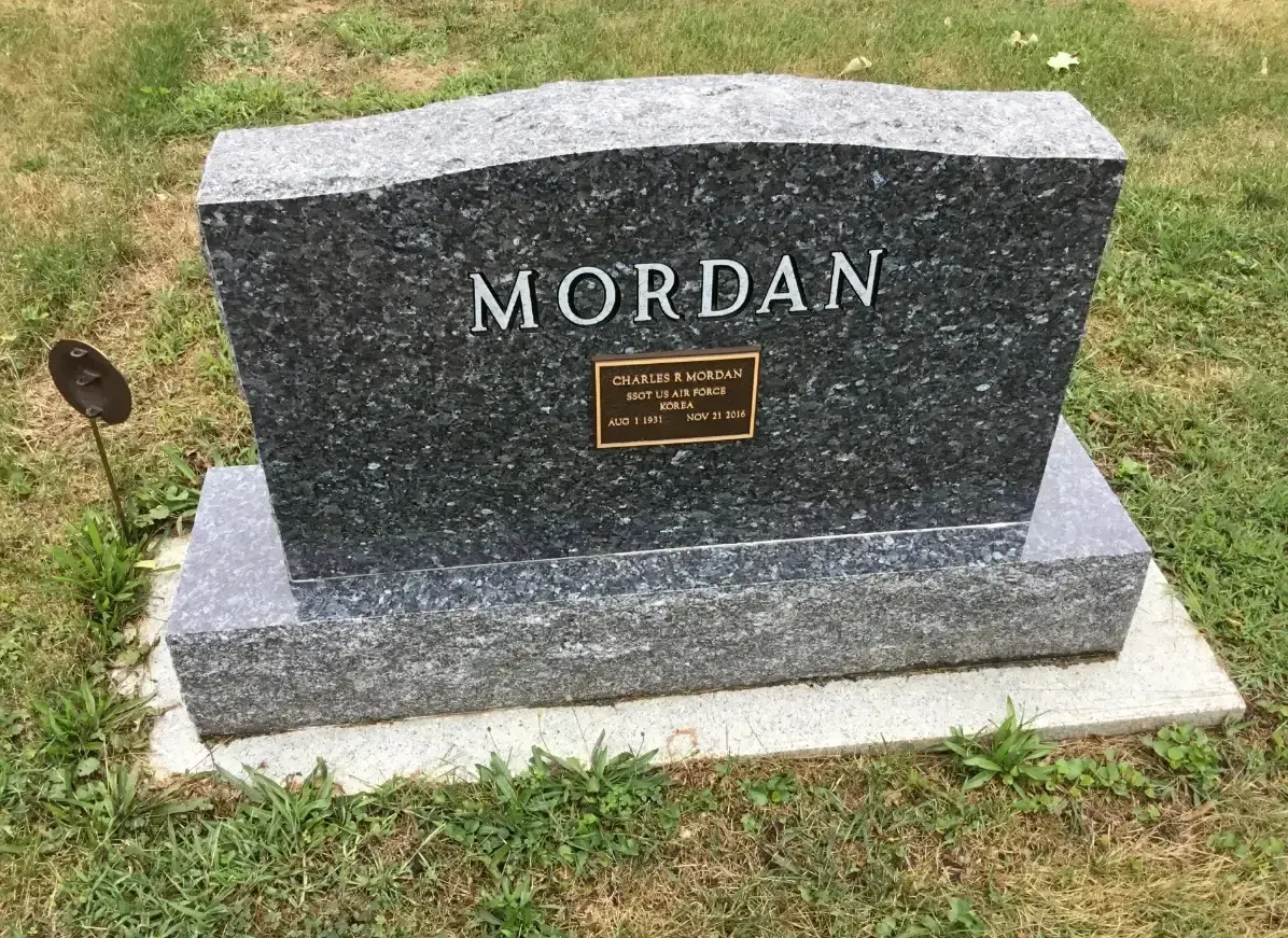 Gravestone engraved “MORDAN” with a small plaque, set on a grassy lawn.