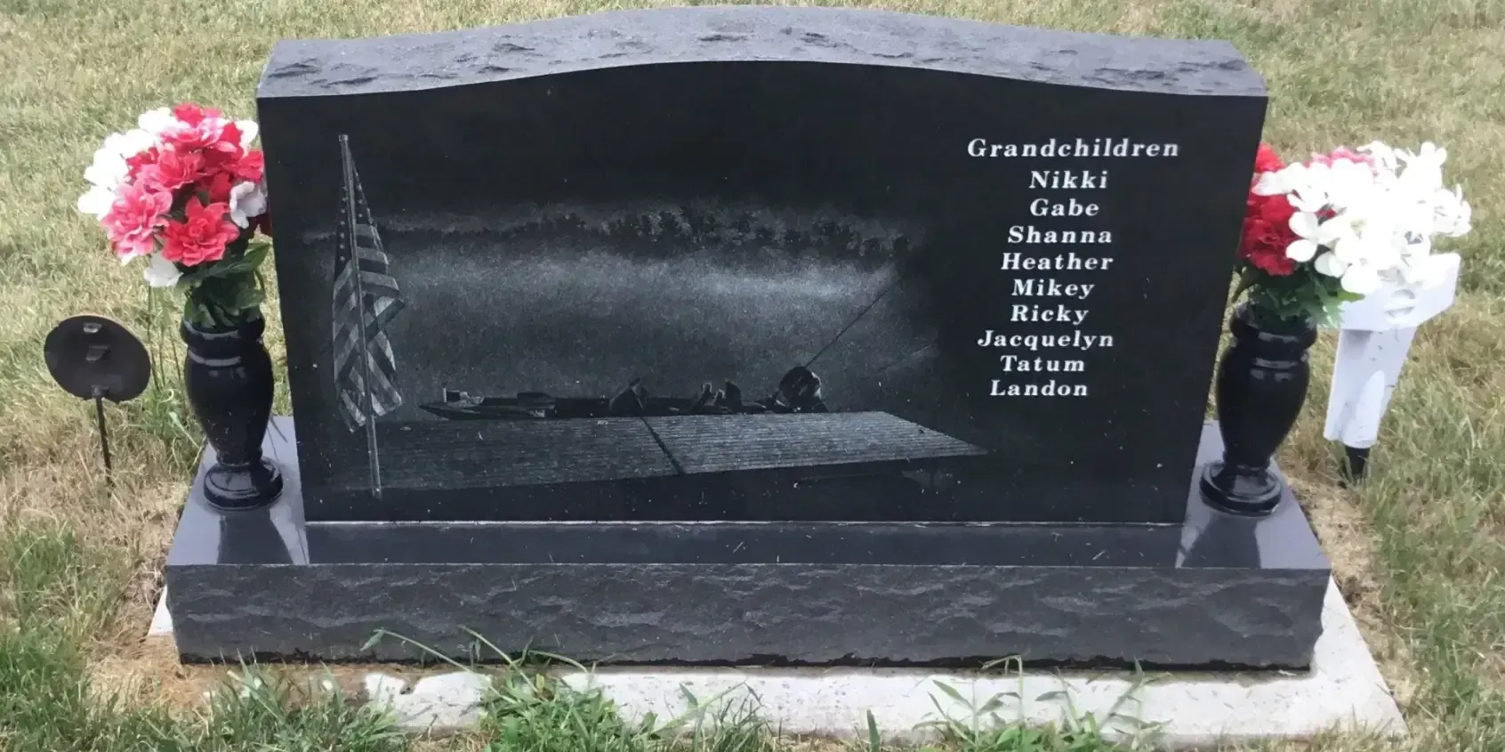 Black granite gravestone with engraved names, flanked by pink and white floral arrangements.