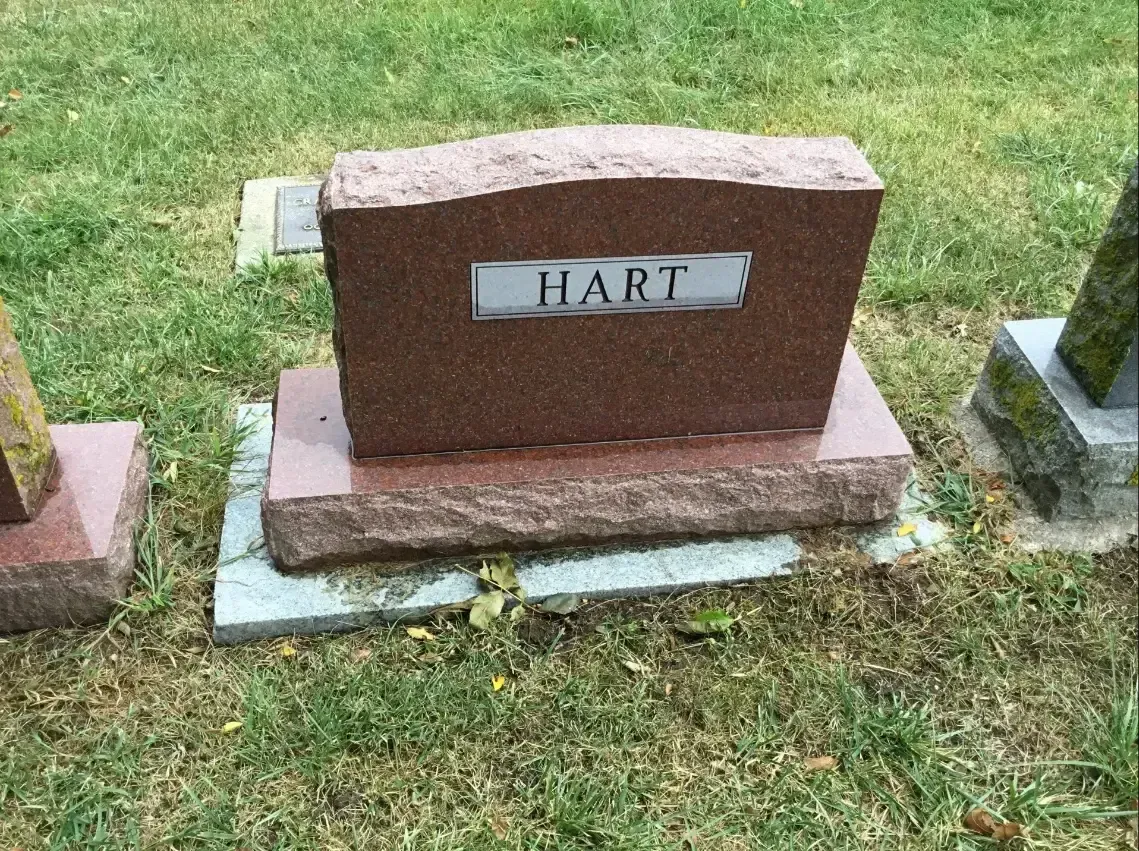Red granite gravestone on grass with a metal plaque reading “HART”