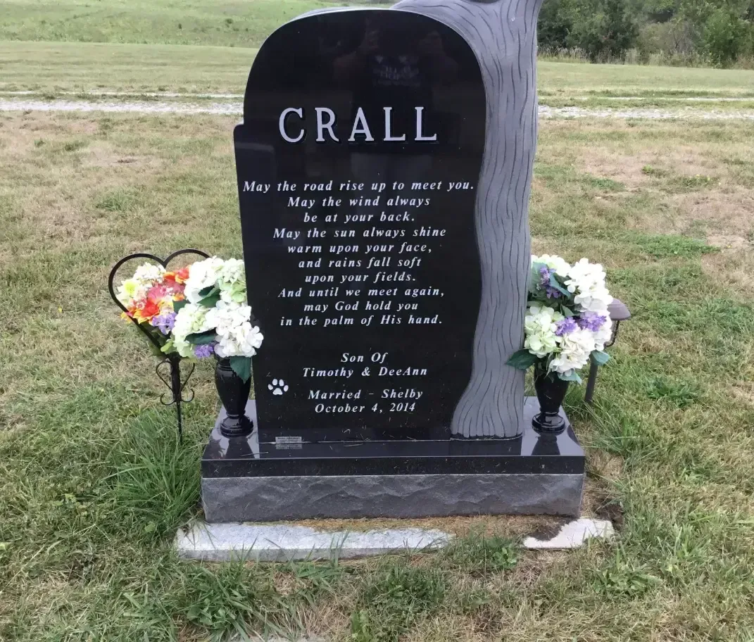 Black Crall gravestone with flowers in a grassy cemetery.