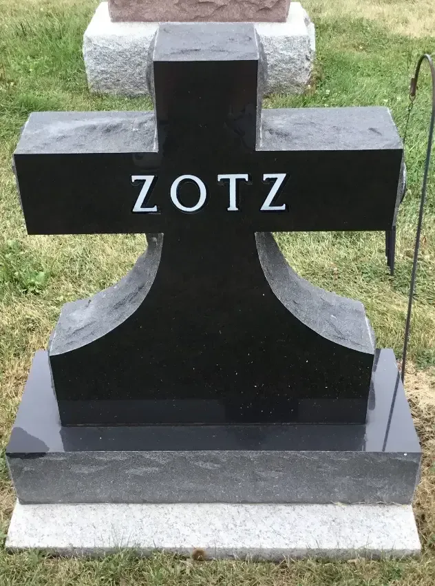 Black gravestone with white letters “ZOTZ” on a grassy lawn, mounted on a gray base