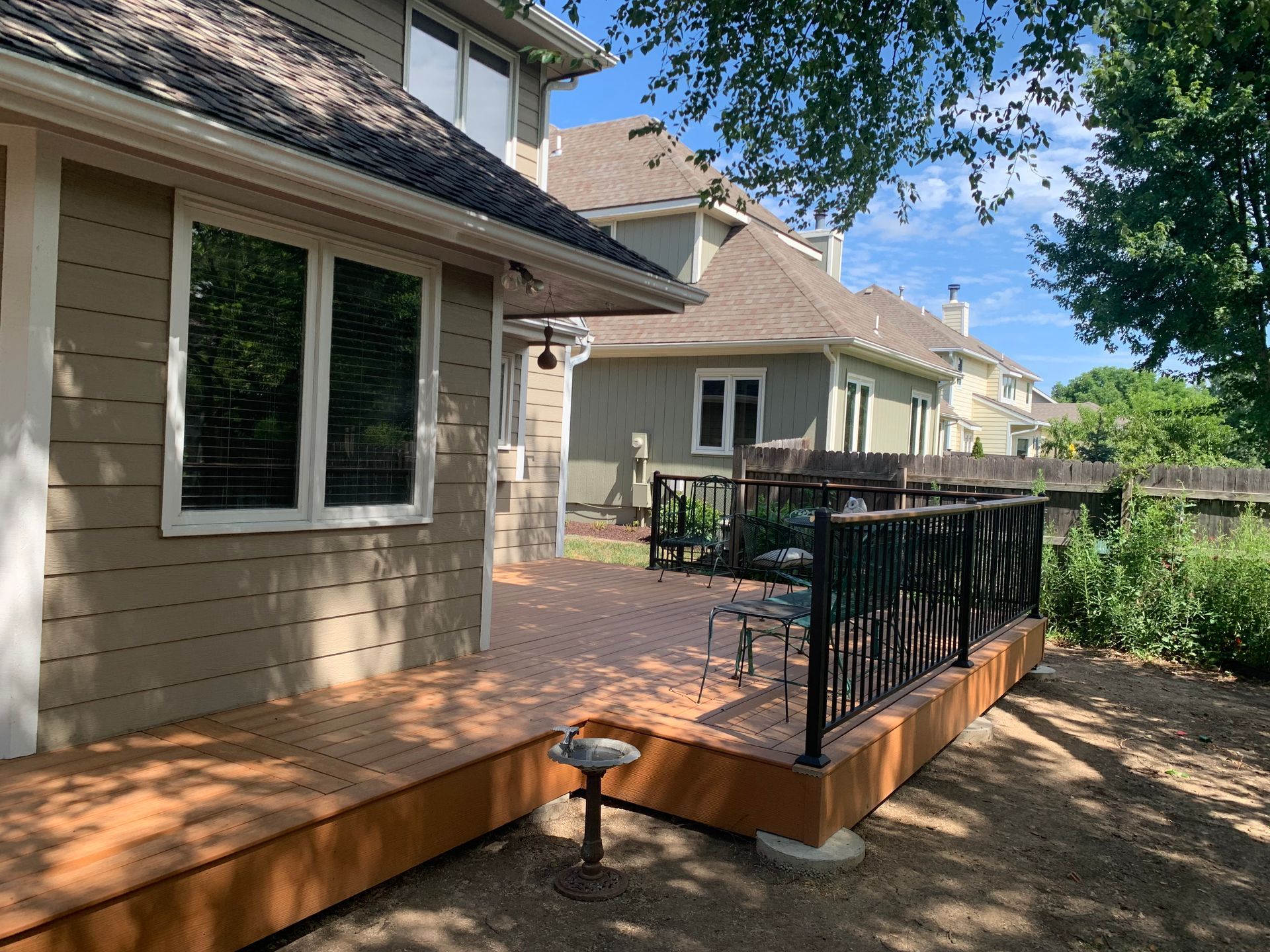 Backyard wooden deck beside a beige house, with black railing and steps leading to a patio.