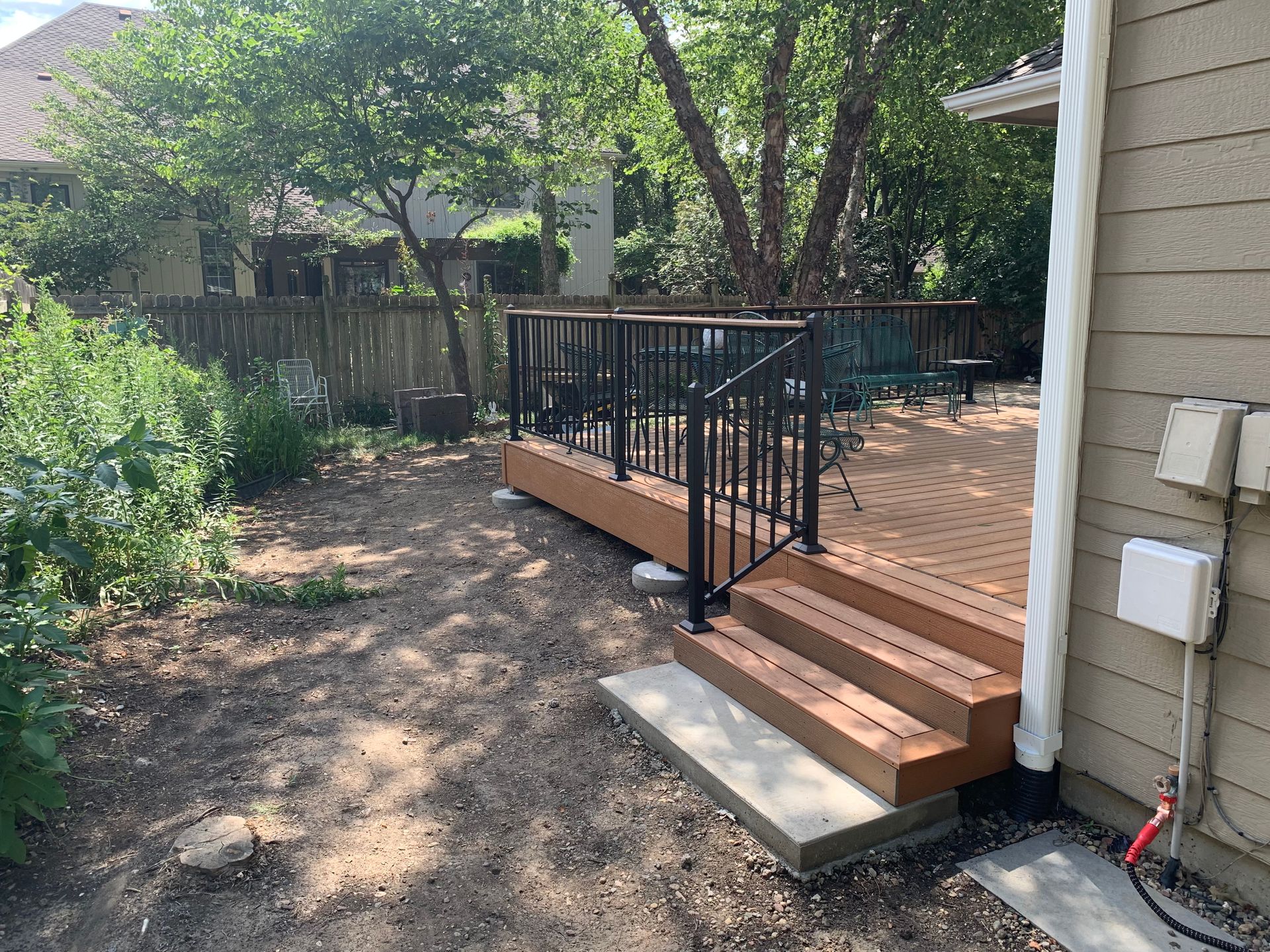 Wooden deck steps with black railing leading to a backyard patio beside a house and trees