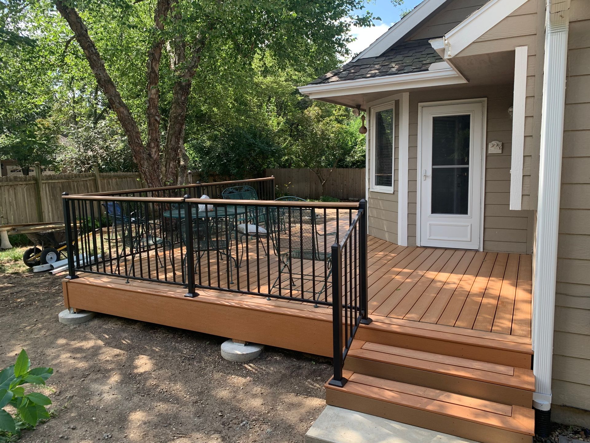 Backyard wooden deck with black railings and steps beside a house, surrounded by trees and dirt ground