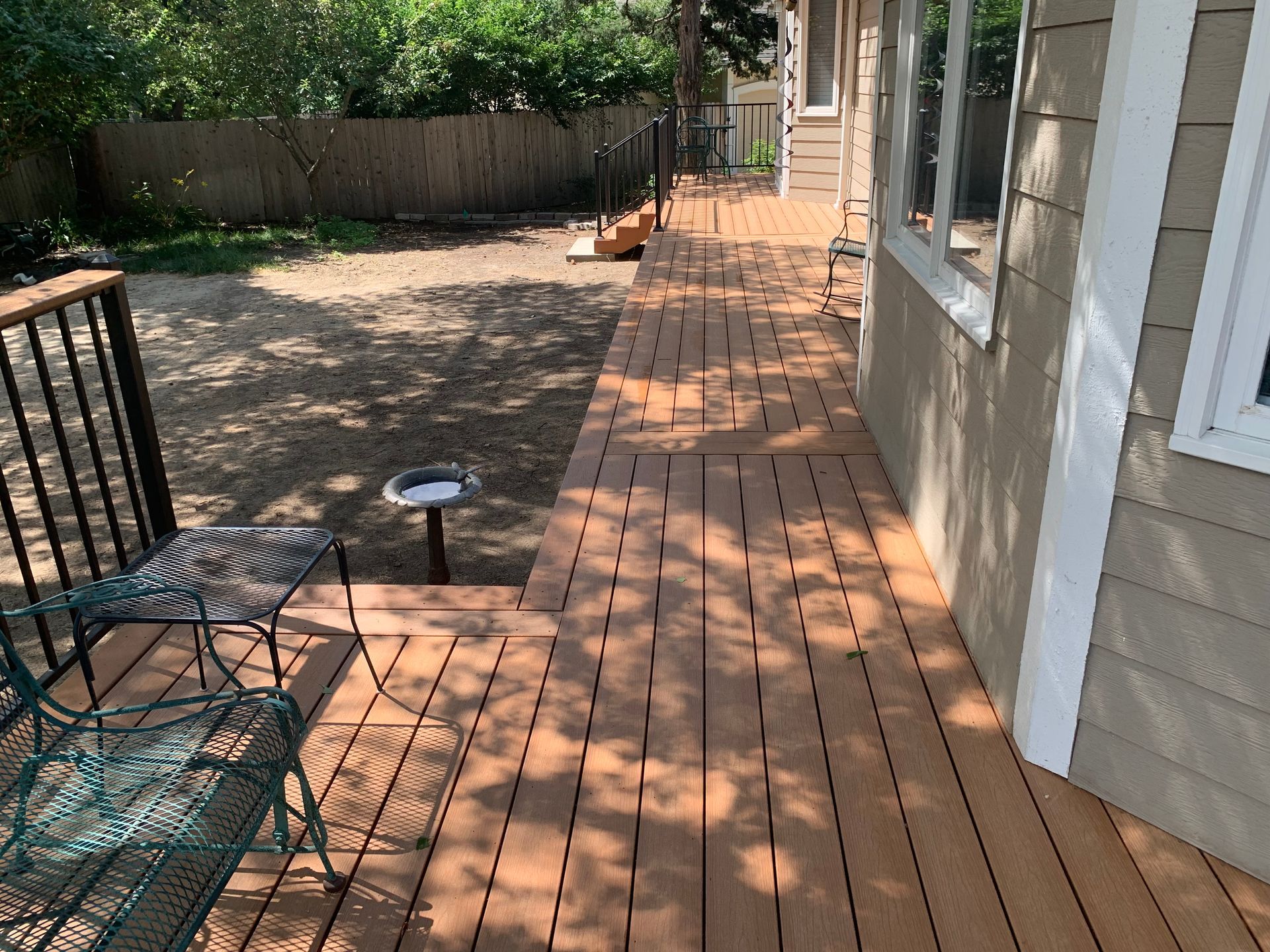 Wooden backyard deck beside a house, with patio chairs, a small table, and a fenced yard in dappled sunlight.