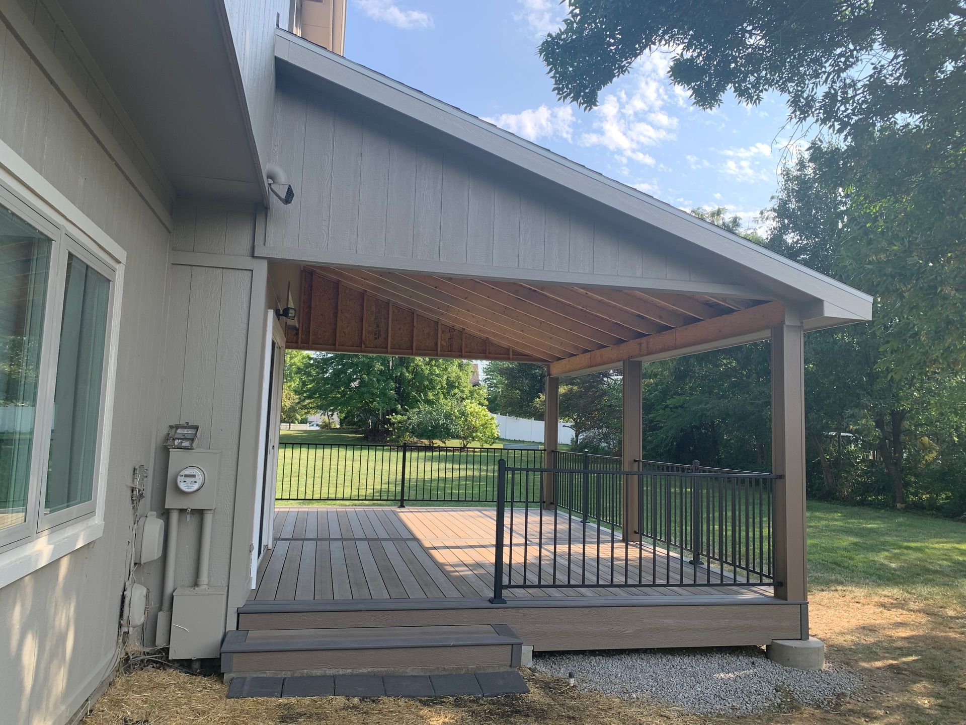 Covered wooden deck and patio with railing beside a gray house, overlooking a grassy yard.
