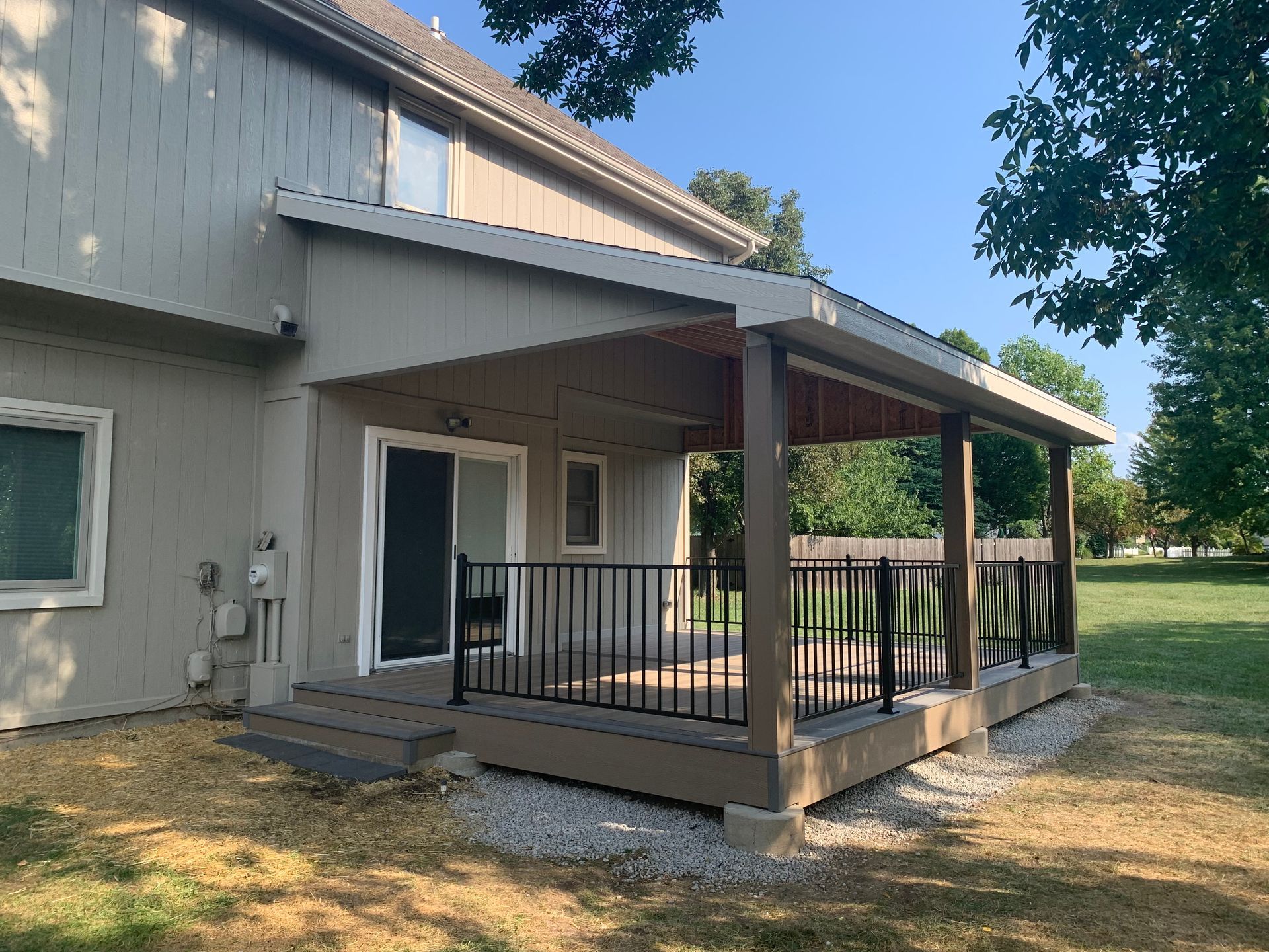 Gray house with a covered wooden patio and black railing beside a grassy yard