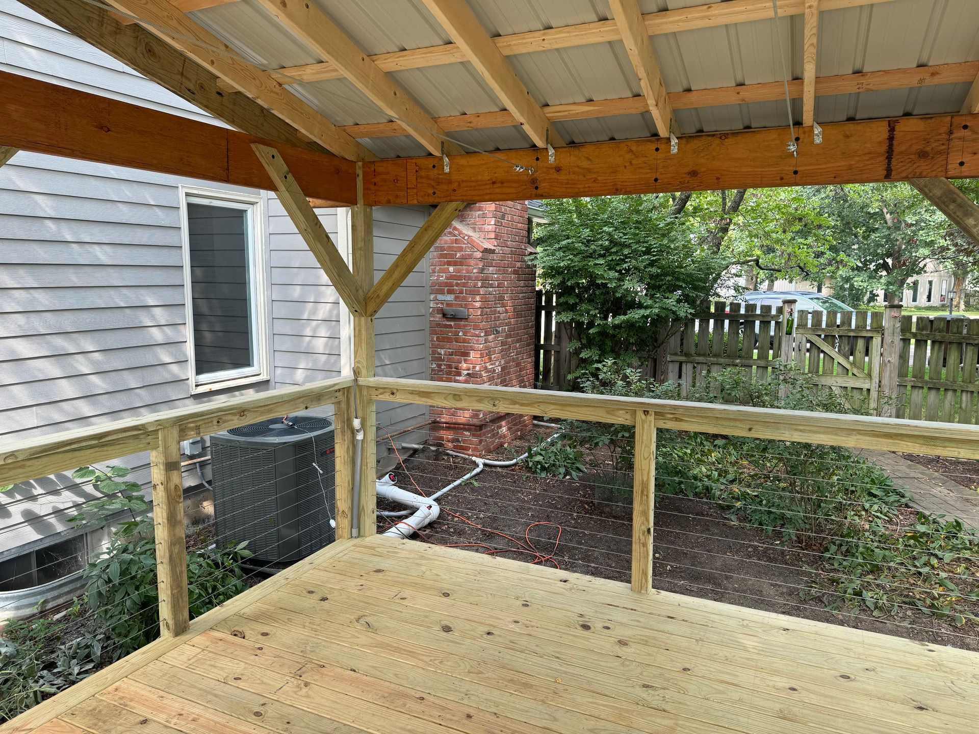 Covered wooden deck with railing beside a house and fenced backyard, viewed from under the roof