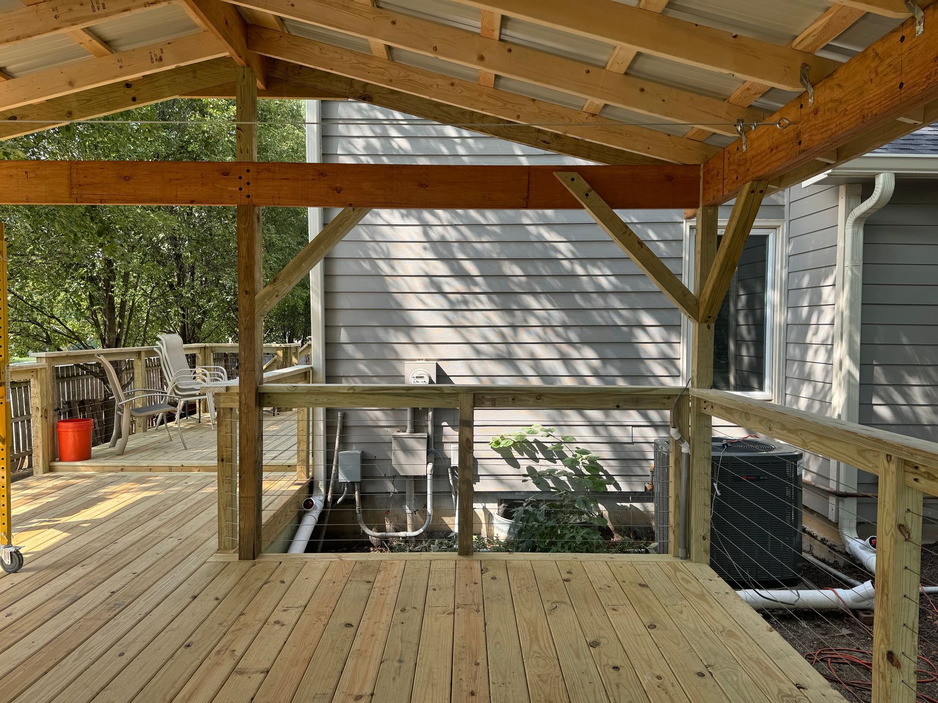 Covered wooden deck with exposed beams and stairs beside a house, overlooking a yard.