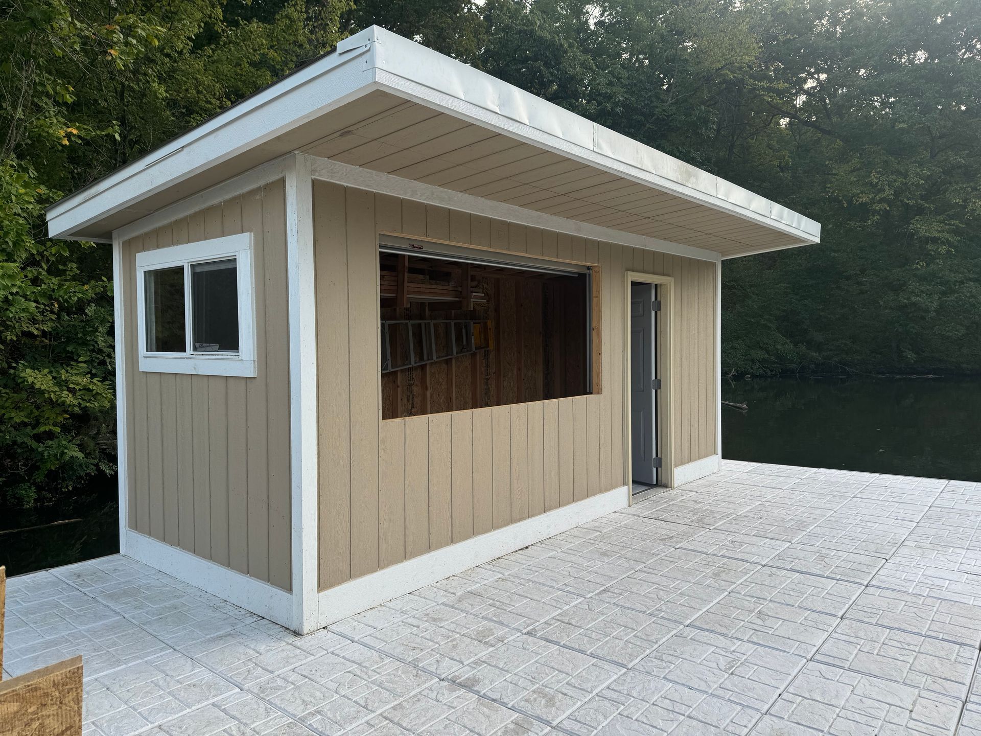 Small beige kiosk with white trim on a paved waterfront deck, backed by water and trees.