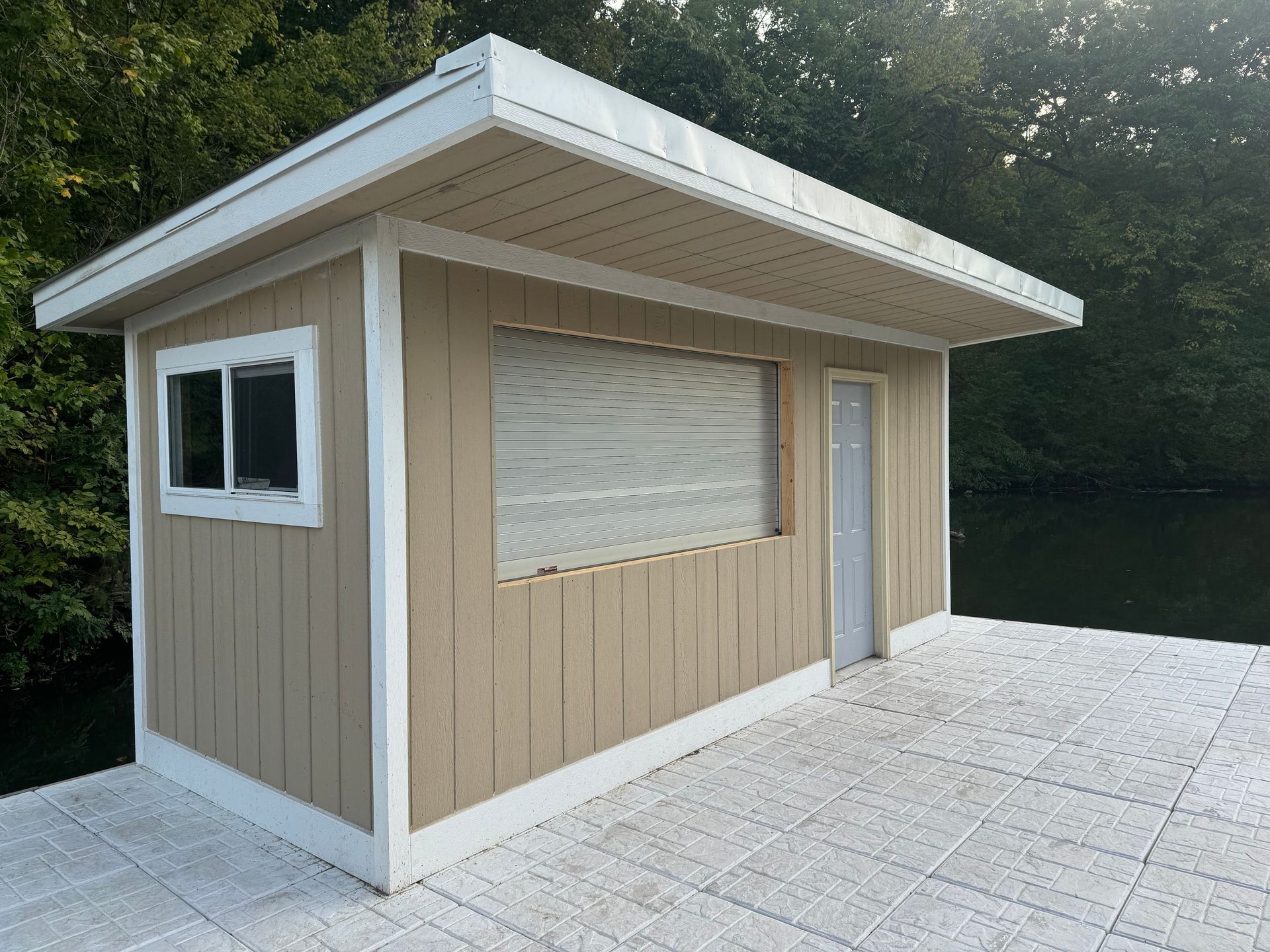 Small tan-and-white utility building with shuttered window and door on a paved patio beside trees