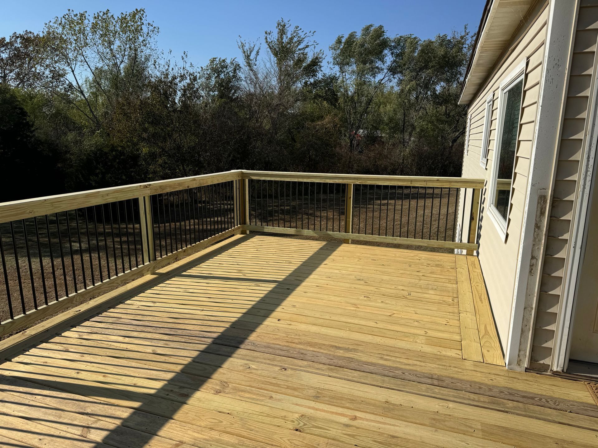 Wooden deck with railing beside a house, overlooking trees in bright sunlight.