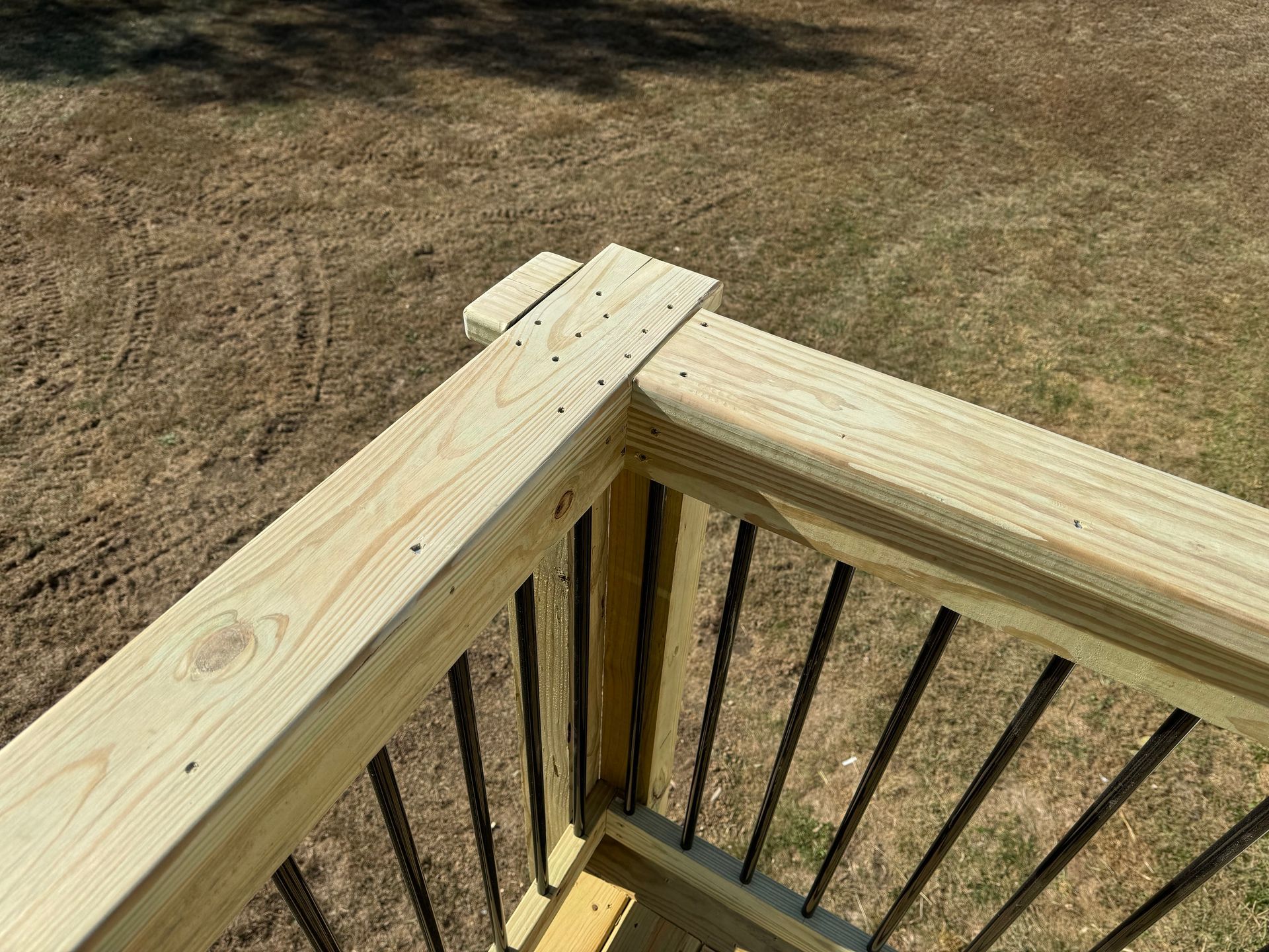 Wooden deck railing corner with black balusters overlooking a dry grassy yard