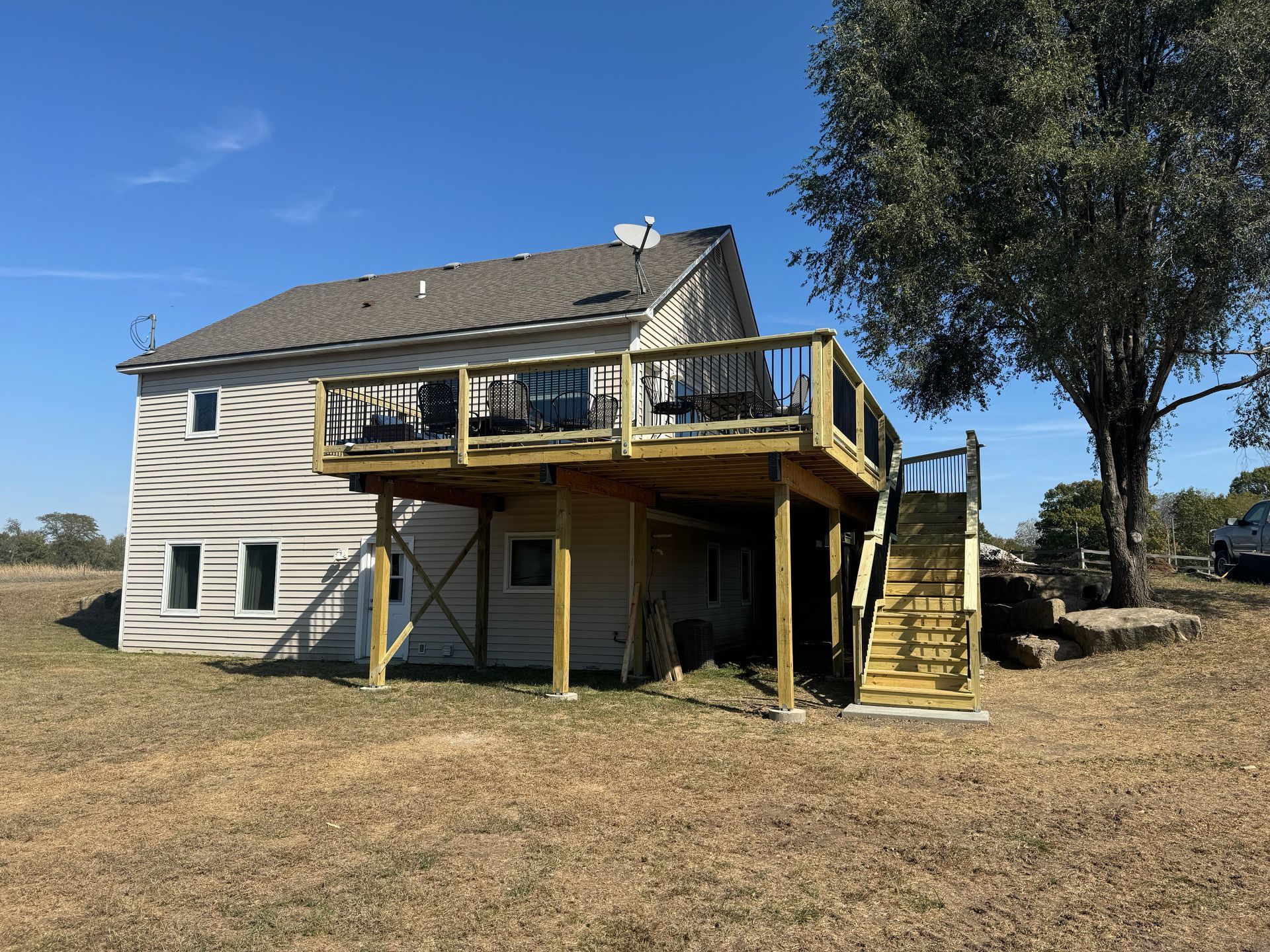Back of a beige house with a raised wooden deck and stairs in a grassy yard under a blue sky