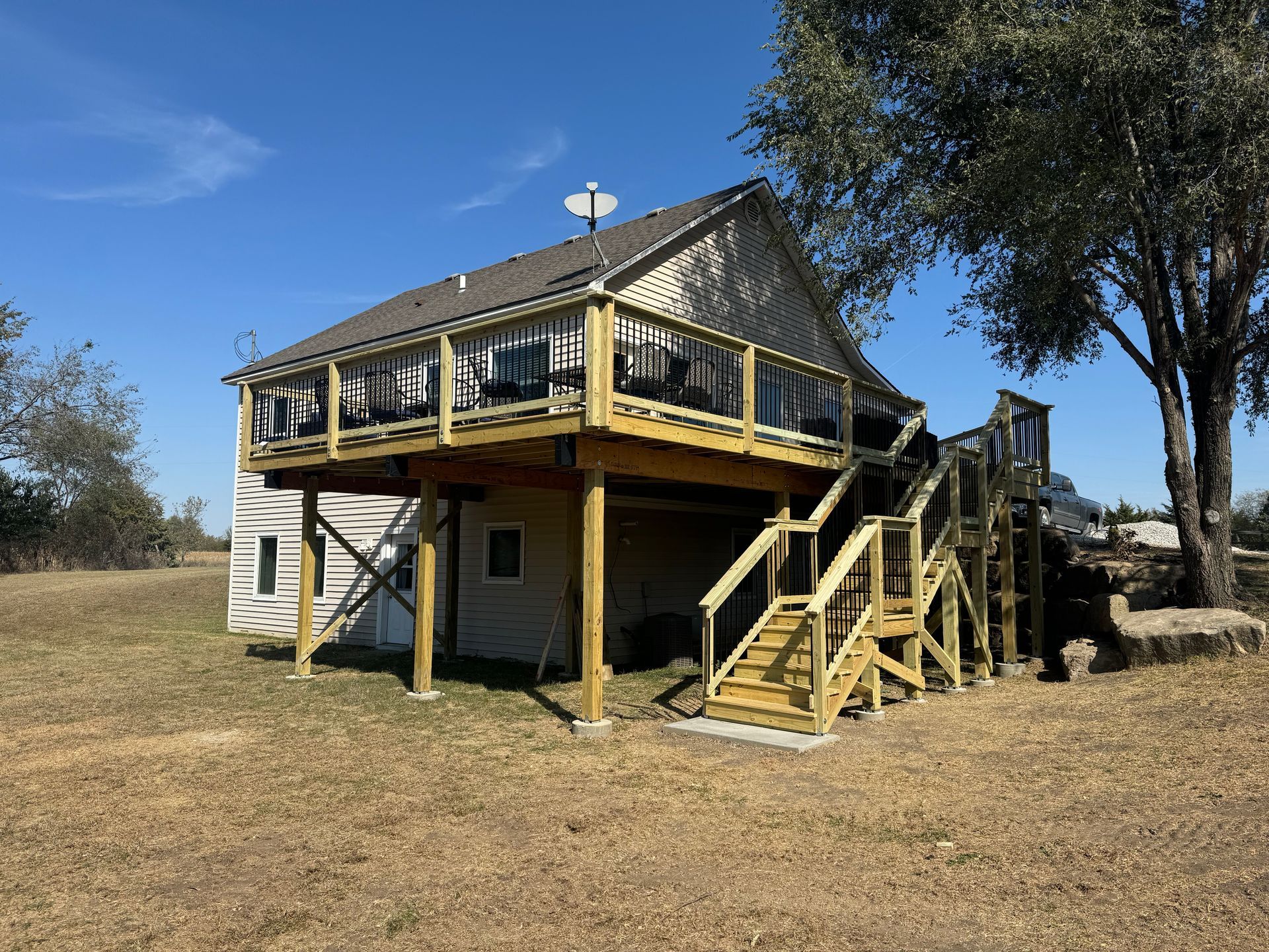 Two-story wooden house with a wraparound deck and exterior stairs in a dry grassy yard under a blue sky