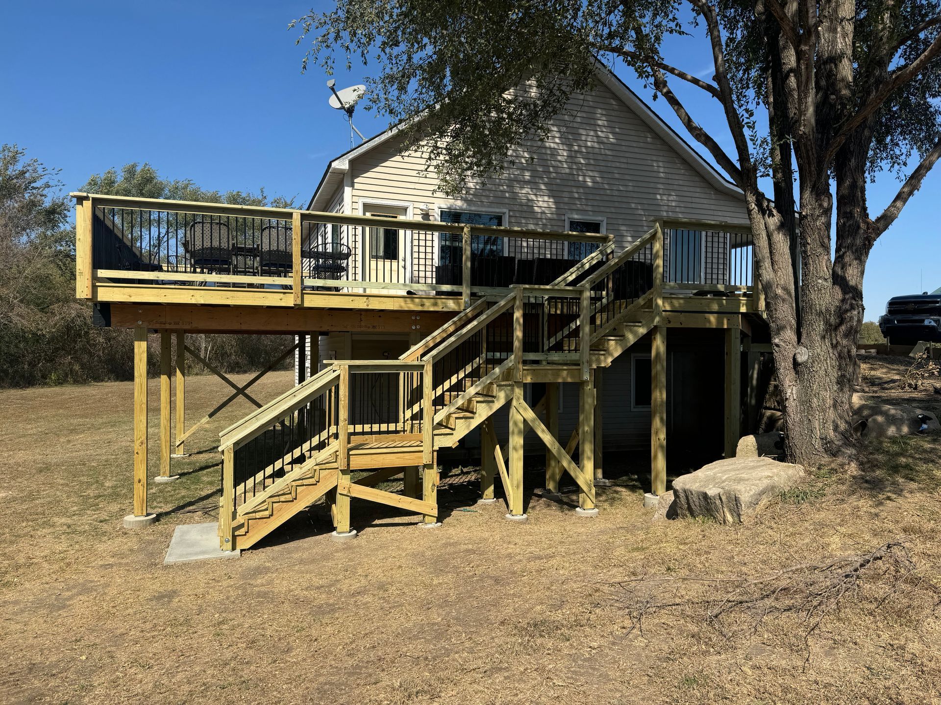 Wooden house with large wraparound deck and stairs in a dry grassy yard beneath trees