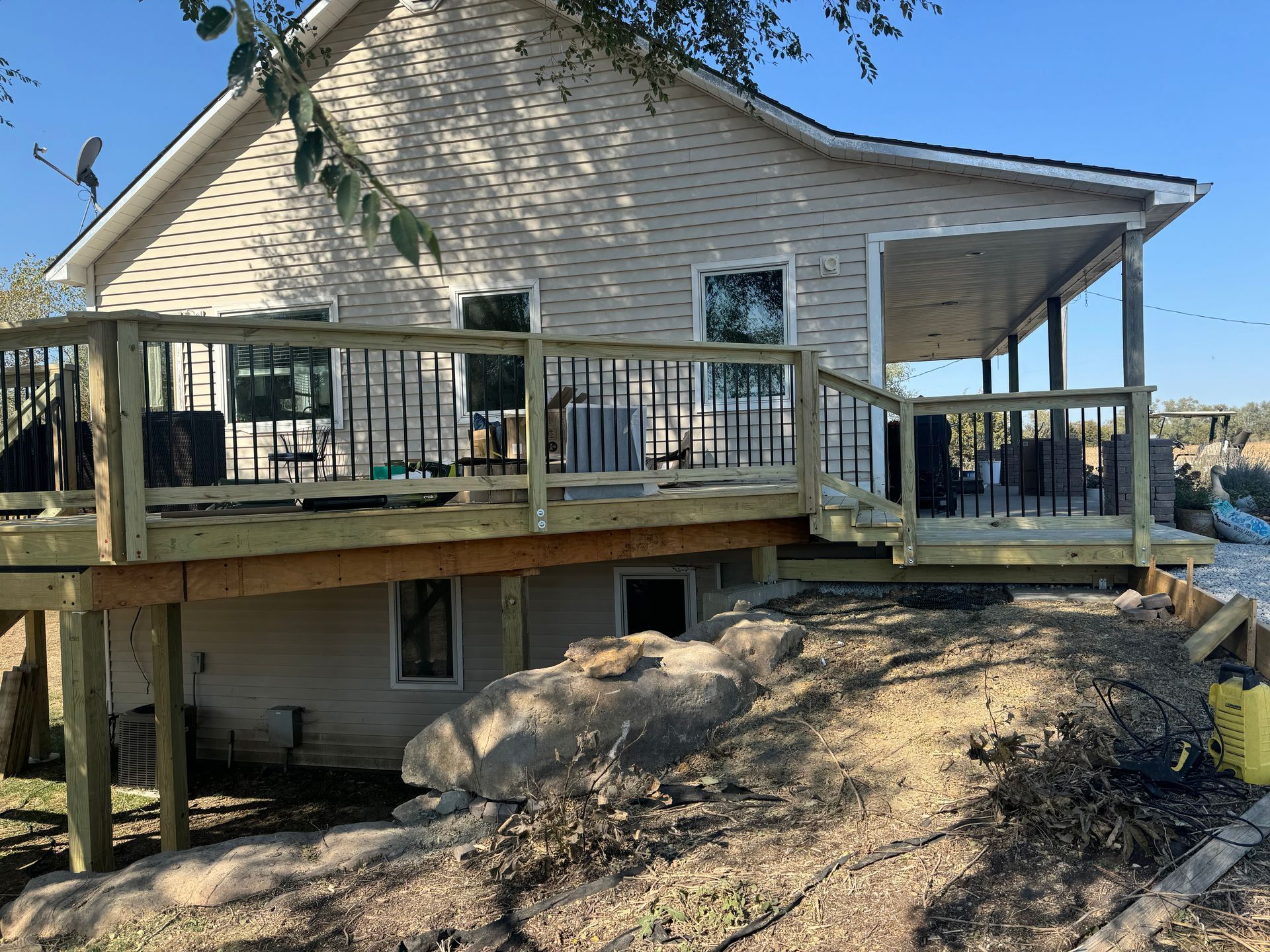 Raised wooden deck on a house beside a large boulder, with stairs and a roofed porch under clear sky