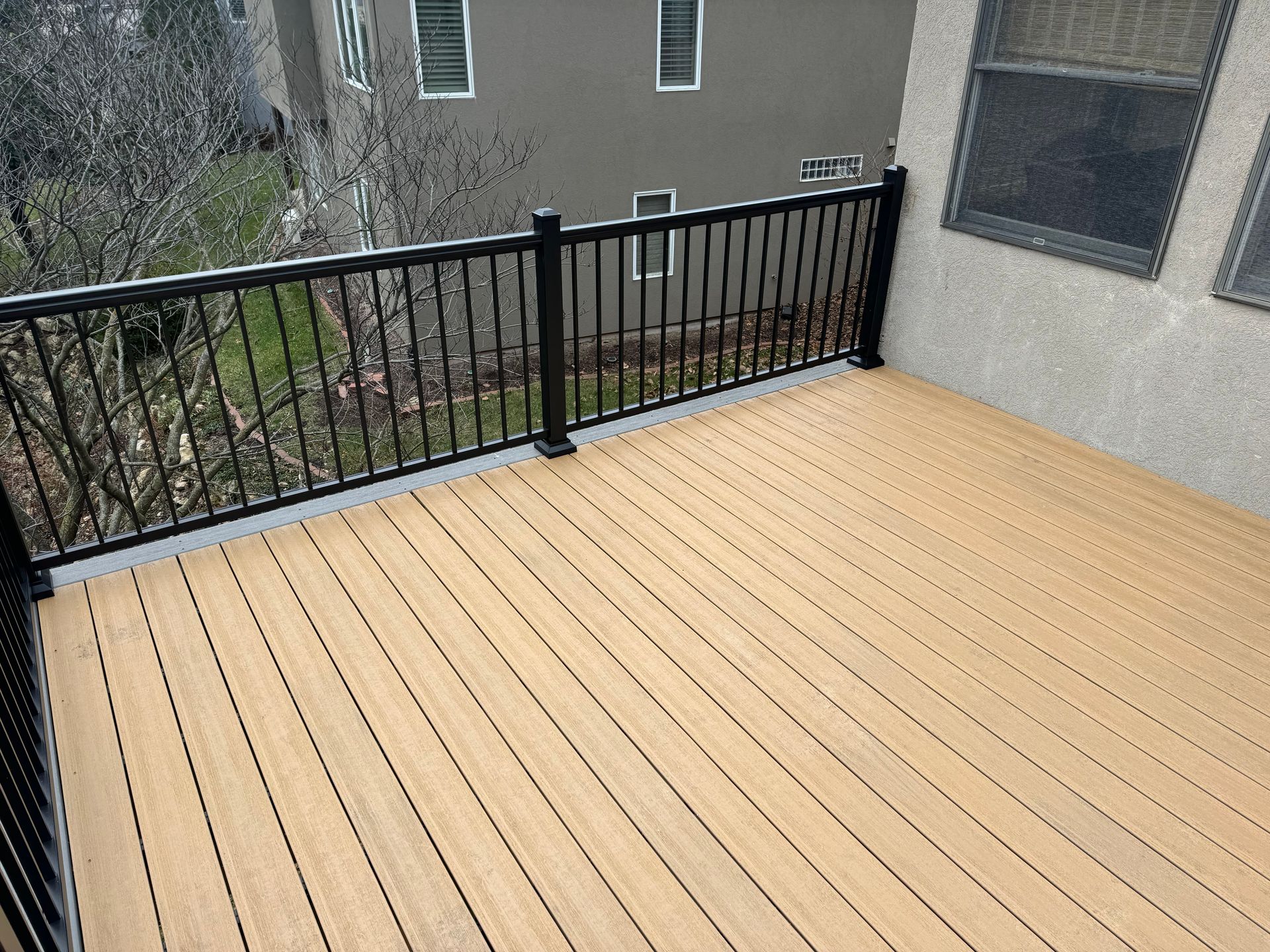 Empty wooden deck with black railing beside a stucco building, overlooking trees and neighboring apartments