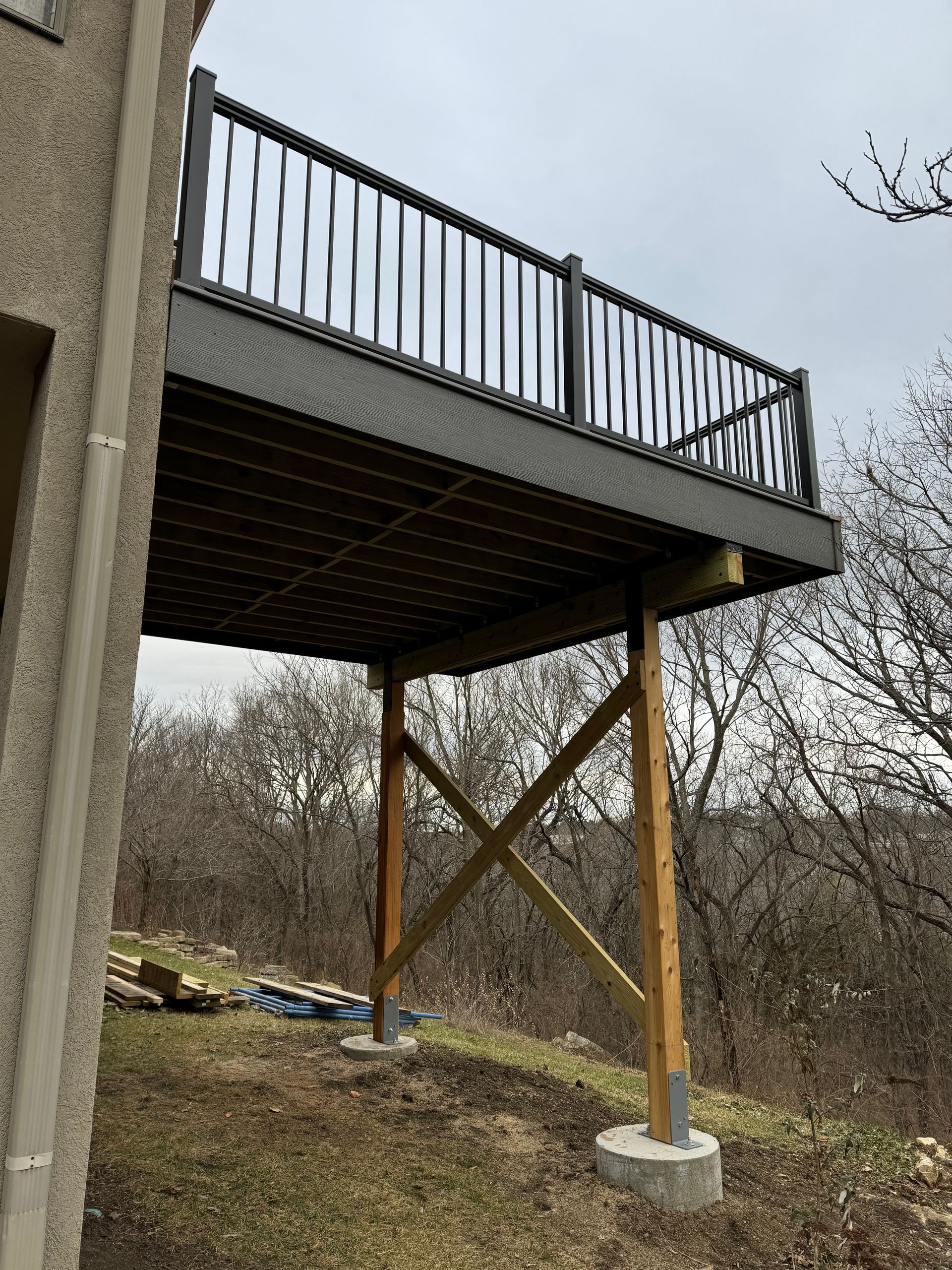 Raised deck with black railing, supported by wooden posts over a sloped yard in winter.
