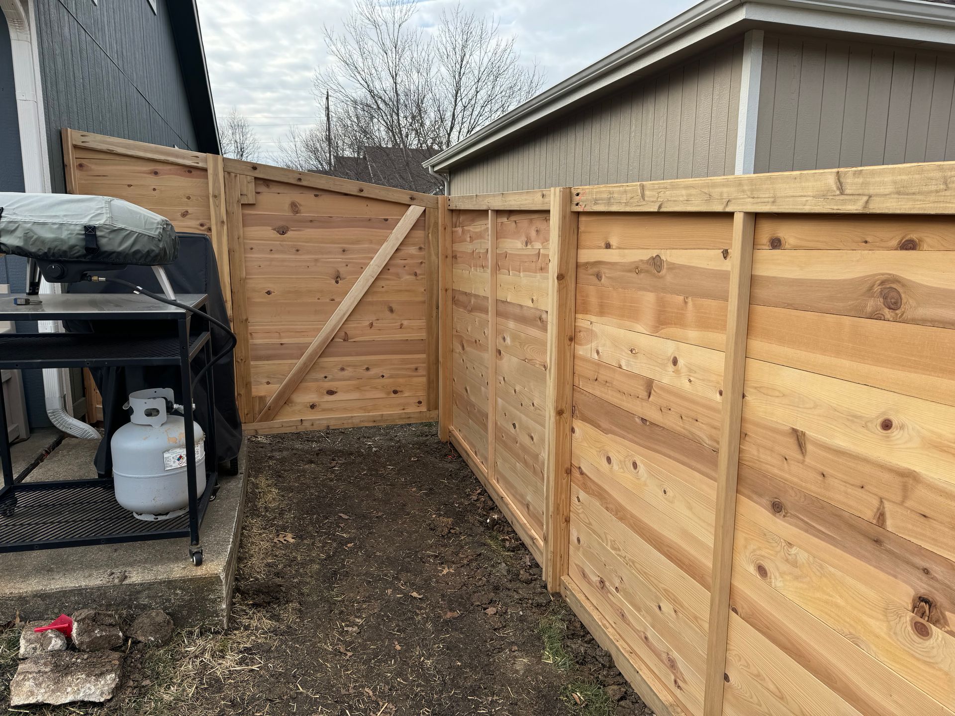 Narrow gravel side yard with a wooden privacy fence and a covered grill on the left.