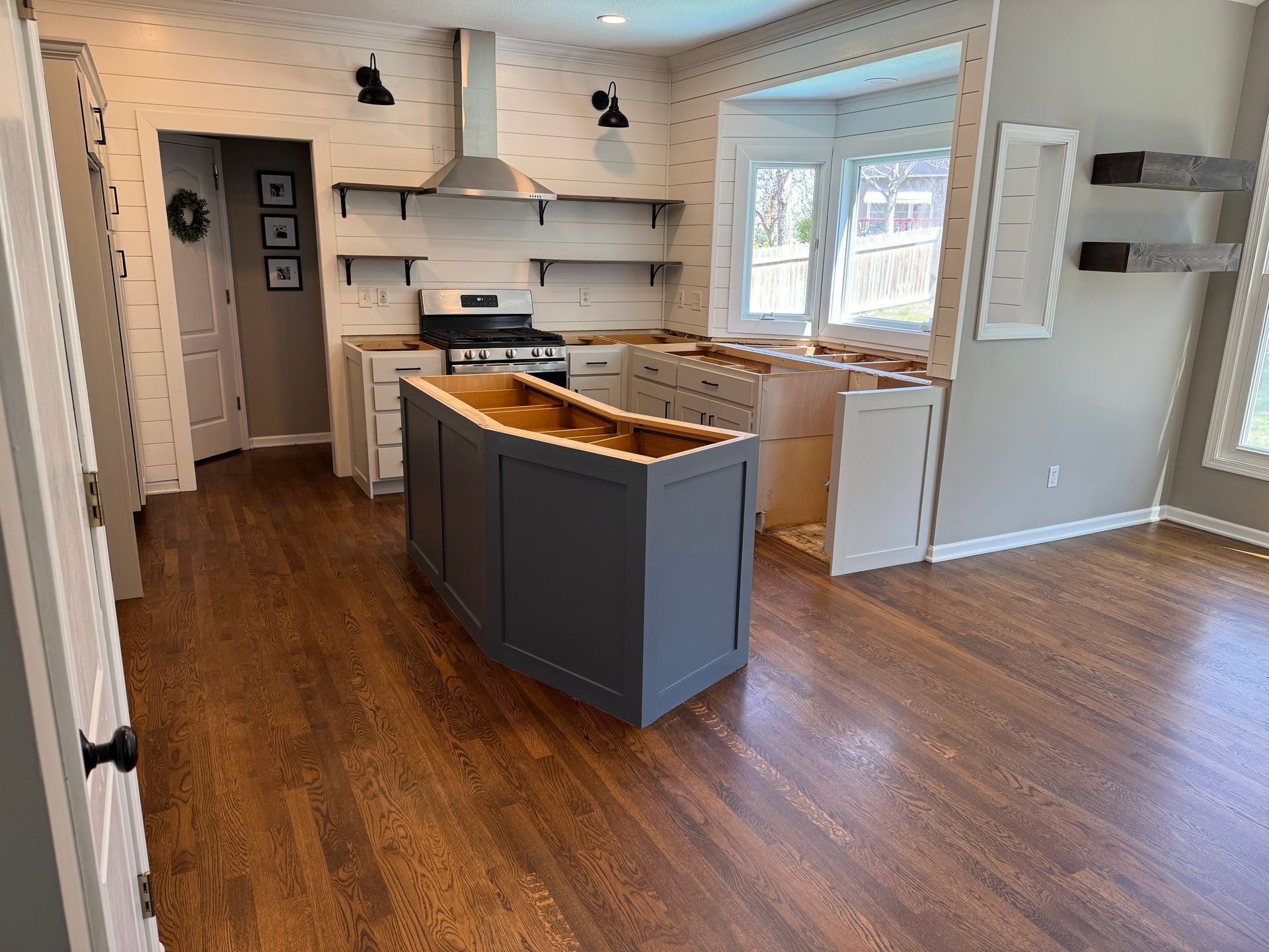 Bright, empty kitchen with wood floors, gray island, white cabinets, and large window.
