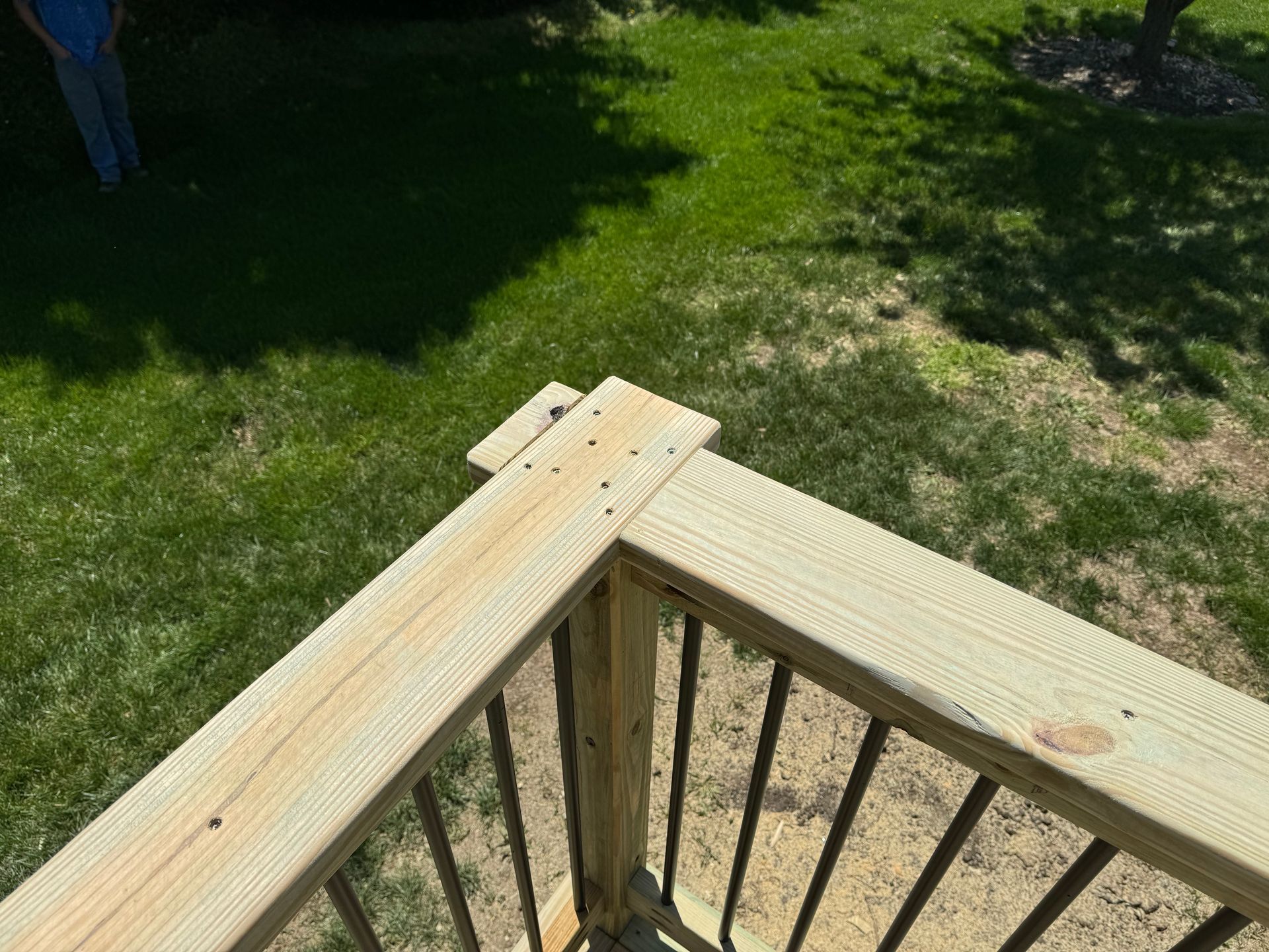 Wooden deck corner with metal balusters overlooking a green lawn and shaded yard.