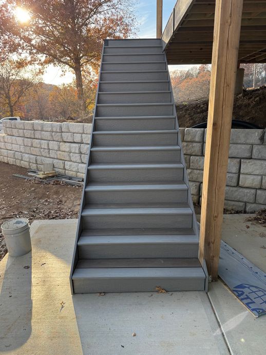 Outdoor concrete staircase beside a stone wall and building, with sunlit trees in the background.